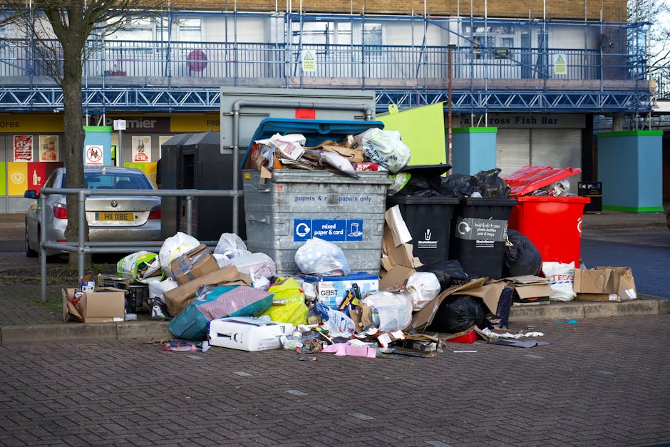 A collection of overflowing rubbish bins and numerous scattered waste items occupy the curbside of a paved street in front of a commercial building. The waste includes various cardboard boxes, plastic bags, paper, and other refuse, some of which pile up around the bins and spill onto the pavement. The central grey recycling bin, labeled for mixed paper and cardboard, is topped with loose rubbish, with additional black and red waste containers adjacent. Behind the trash, there is a parked silver car with a yellow license plate nearby, and a metal railing separates the waste area from the street. In the background, the building features a shopfront with a bright yellow sign and a blue scaffolding structure covering parts of the upper facade. The scene is illuminated by natural daylight, highlighting the disorderly state of the disposal area, which relates to private waste collection methods often used as alternatives to municipal rubbish services, aligning subtly with the theme of rubbish removal services provided by Waste Disposal Brompton.
