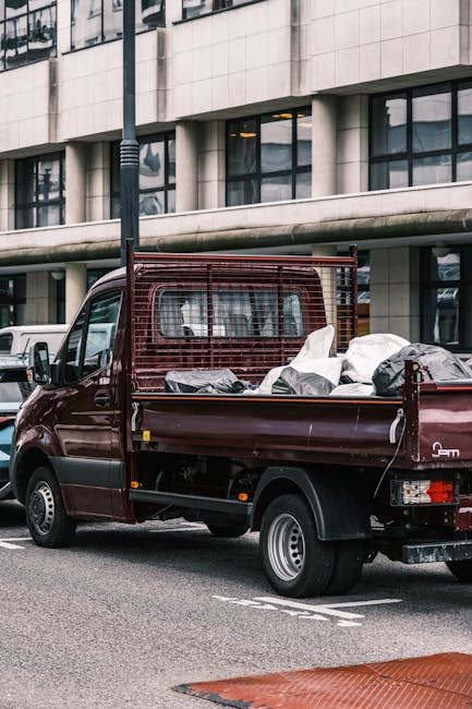 A burgundy flatbed van parked on an urban street with a modern office building in the background, featuring large glass windows and a concrete facade. The vehicle is loaded with various waste materials, including several black and white bags, some appearing to be filled with household or construction debris. The cargo area has a metal grid on the sides and rear, providing support and containment for the waste items. The van’s rear lights are visible, and it is positioned next to a painted parking space. The scene is lit by natural daylight, with a streetlamp visible near the vehicle. This image relates to private waste collection services, such as those offered by Waste Disposal Brompton, which provide alternative rubbish removal options outside of local authority collection, emphasizing the accessibility of on-site clearance for household or commercial waste disposal needs.