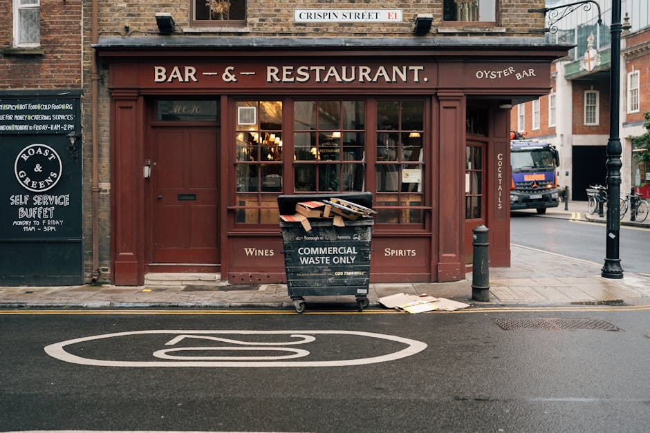 In front of a brick-fronted bar and restaurant with a deep red wooden exterior, a large black commercial waste container labeled 'COMMERCIAL WASTE ONLY' is positioned on the street curb, filled with flattened cardboard boxes and paper packaging, some partially protruding from the top. The container is situated between a black bollard and a pole, with discarded paper sheets lying on the pavement nearby. The restaurant's windows reveal warm interior lighting, and signage indicates that the establishment serves wines, spirits, and cocktails. To the left, a blackboard-style sign advertises a self-service buffet, and on the right side, a black waste truck is partially visible behind the storefront. The scene is set on a typical city street with a designated no-parking road marking painted directly on the tarmac, and the environment suggests an urban setting that may be serviced by private waste removal companies like Waste Disposal Brompton, providing an alternative to traditional council rubbish collection for local businesses.