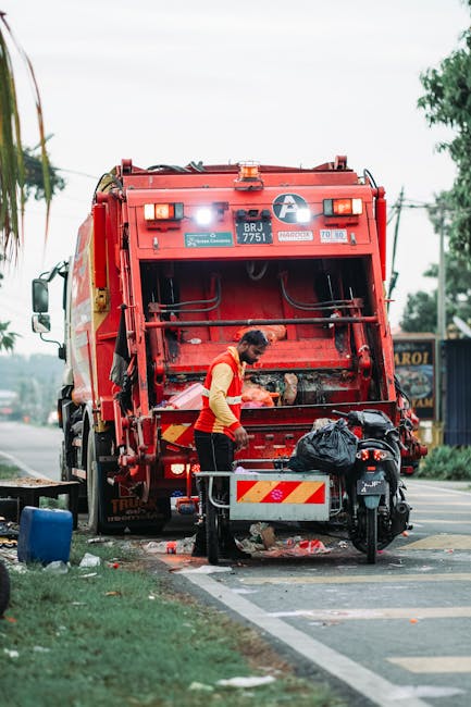 A sanitation worker is seen operating a red rubbish collection truck on the side of a street, with its rear compartment open and filled with waste. The worker, dressed in a high-visibility orange vest over dark clothing, is standing next to a metal collection bin and appears to be managing waste materials. The truck's rear features reflective hazard markings, identification labels, and a hydraulic system for loading rubbish. A black garbage bag and a motorcycle are parked nearby on the curb. The scene takes place in a suburban or urban environment, with trees, power lines, and a commercial sign visible in the background. The image is taken during natural daylight, highlighting the functional and practical elements of private waste collection, consistent with independent rubbish removal services such as those offered by Waste Disposal Brompton.