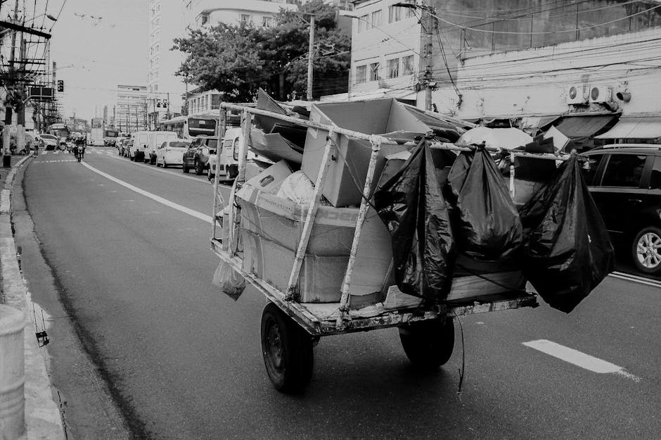 In front of a brick-fronted bar and restaurant with a deep red wooden exterior, a large black commercial waste container labeled 'COMMERCIAL WASTE ONLY' is positioned on the street curb, filled with flattened cardboard boxes and paper packaging, some partially protruding from the top. The container is situated between a black bollard and a pole, with discarded paper sheets lying on the pavement nearby. The restaurant's windows reveal warm interior lighting, and signage indicates that the establishment serves wines, spirits, and cocktails. To the left, a blackboard-style sign advertises a self-service buffet, and on the right side, a black waste truck is partially visible behind the storefront. The scene is set on a typical city street with a designated no-parking road marking painted directly on the tarmac, and the environment suggests an urban setting that may be serviced by private waste removal companies like Waste Disposal Brompton, providing an alternative to traditional council rubbish collection for local businesses.