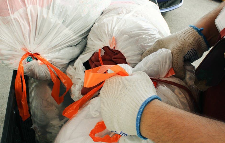 A person wearing beige work gloves with blue trim is handling several large white plastic rubbish bags secured with bright orange plastic ties. The bags appear to contain discarded items and are stacked together, filling a black container or vehicle compartment. In the background, a partial view of a silver car and the pavement can be seen, indicating the scene takes place outdoors, likely in a driveway or parking area. The gloves suggest careful handling typical of waste disposal or rubbish removal tasks, which aligns with private rubbish collection services such as those offered by Waste Disposal Brompton. The scene demonstrates a focused effort on the collection and management of bulky waste, possibly related to an individual or organizational effort to clear excess or non-standard waste materials, supporting alternative rubbish collection options outside of municipal services. The lighting is natural, highlighting the textures of the plastic bags and the gloves, emphasizing a professional and neutral environment dedicated to waste handling activities.