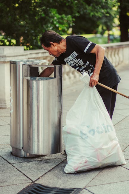 A man with dark hair, wearing a black t-shirt with white text and dark trousers, is seen using a metal trash bin with multiple compartments on a paved outdoor walkway. He is leaning forward and placing or retrieving waste, with his left hand resting on the top edge of the bin. His right hand is holding a large, white plastic rubbish bag, which is partly filled and rests on the ground. The surrounding environment features a stone balustrade and lush green trees in the background, suggesting a park or public space. The scene is well-lit with natural daylight, and the metal bins appear modern and durable, suitable for public rubbish collection. This setup could relate to independent or private waste management services, such as those offered by Waste Disposal Brompton, especially for on-site or alternative rubbish removal in a public outdoor setting.