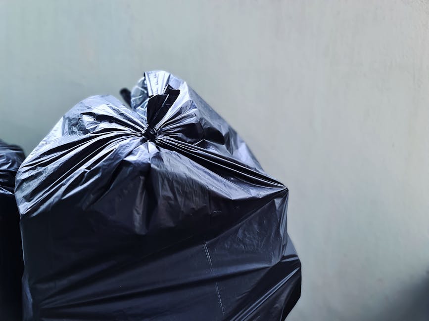 A person wearing beige work gloves with blue trim is handling several large white plastic rubbish bags secured with bright orange plastic ties. The bags appear to contain discarded items and are stacked together, filling a black container or vehicle compartment. In the background, a partial view of a silver car and the pavement can be seen, indicating the scene takes place outdoors, likely in a driveway or parking area. The gloves suggest careful handling typical of waste disposal or rubbish removal tasks, which aligns with private rubbish collection services such as those offered by Waste Disposal Brompton. The scene demonstrates a focused effort on the collection and management of bulky waste, possibly related to an individual or organizational effort to clear excess or non-standard waste materials, supporting alternative rubbish collection options outside of municipal services. The lighting is natural, highlighting the textures of the plastic bags and the gloves, emphasizing a professional and neutral environment dedicated to waste handling activities.