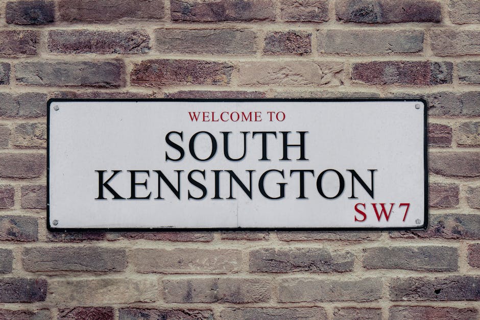 A rectangular white metal street sign mounted on a red brick wall, displaying the words 'Welcome to South Kensington' in bold black lettering, with 'SW7' in smaller red font at the bottom right corner. The sign has a slightly textured surface with visible screws at each corner securing it to the wall. The brick wall behind exhibits a traditional pattern with varying shades of red and brown, providing a textured background. The setting suggests an urban environment, possibly near a station or residential area, where private waste clearance services like those offered by Waste Disposal Brompton may operate, supporting alternative rubbish removal methods aside from local authority collection.
