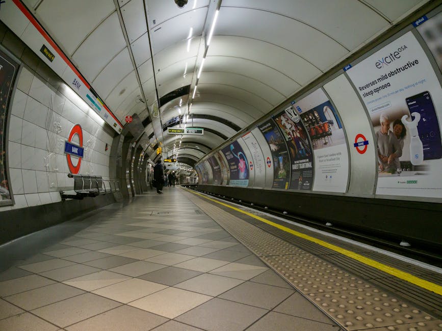 An underground railway station platform with a curved ceiling lined with white tiles and illuminated by long, narrow fluorescent lights. On the left, there's a row of metal benches and a red and blue London Underground roundel sign mounted on the wall. The platform surface is paved with beige and gray tiles arranged in a diagonal pattern, with tactile paving strips along the edge near the tracks. To the right, a continuous advertising panel displays various posters and promotional content, including an advertisement for a sleep device, accompanied by the London Underground logo. A few passengers are visible standing or walking in the distance, with the tracks and a queueing area extending further down the platform. The overall atmosphere reflects a typical modern tube station interior, designed for passenger safety and information dissemination, with an emphasis on functional lighting and signage for public transit and associated services.