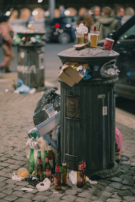 A black, cylindrical public waste bin with a domed lid, located on a cobbled street in an urban area. The bin is covered with various discarded items, including used paper cups with plastic lids and straws, some with remnants of beverage inside, and a crumpled red carton. The side of the bin has an open slot with a partially filled plastic bag of waste hanging from it. Surrounding the bin on the ground are several glass bottles, some with liquid inside, and a black plastic garbage bag tied at the top, along with scattered rubbish such as paper and plastic wrappers. In the background, pedestrians are walking, and parked cars are visible, indicating a busy street setting. The scene suggests a typical example of street littering demonstrating the importance of proper rubbish disposal, relevant to waste removal services provided by Waste Disposal Brompton, especially in urban environments where occasional improper waste management occurs.