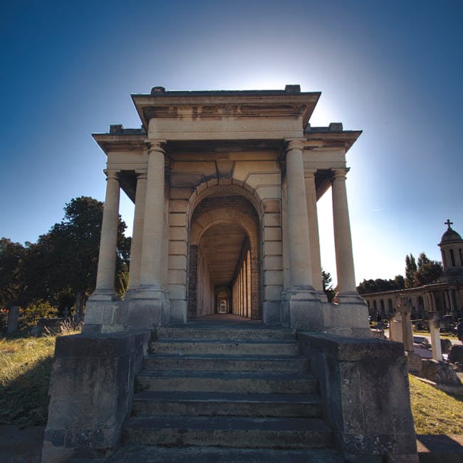 A historic stone mausoleum featuring a neoclassical architectural style with tall, smooth, cream-colored columns supporting a rectangular entablature and a flat roof. The mausoleum is accessed via a short set of stone steps leading up to an archway passageway, which extends through the structure, revealing a corridor lined with columns on either side. The stone surface has a weathered and slightly textured appearance, with some darker patches indicating age and exposure. Surrounding the mausoleum is a manicured grassy area with a few trees visible on either side, bathed in bright natural sunlight that creates a vivid backdrop with a clear blue sky. Other mausoleums and gravestones are visible in the background, indicating a cemetery setting. The scene is quiet and well-maintained, reflecting a traditional burial site, with no presence of waste or rubbish visible. Waste Disposal Brompton’s approach to private waste management might be relevant for maintaining such sites, although the image itself depicts a clean and orderly environment suitable for professional rubbish removal and on-site clearance services.